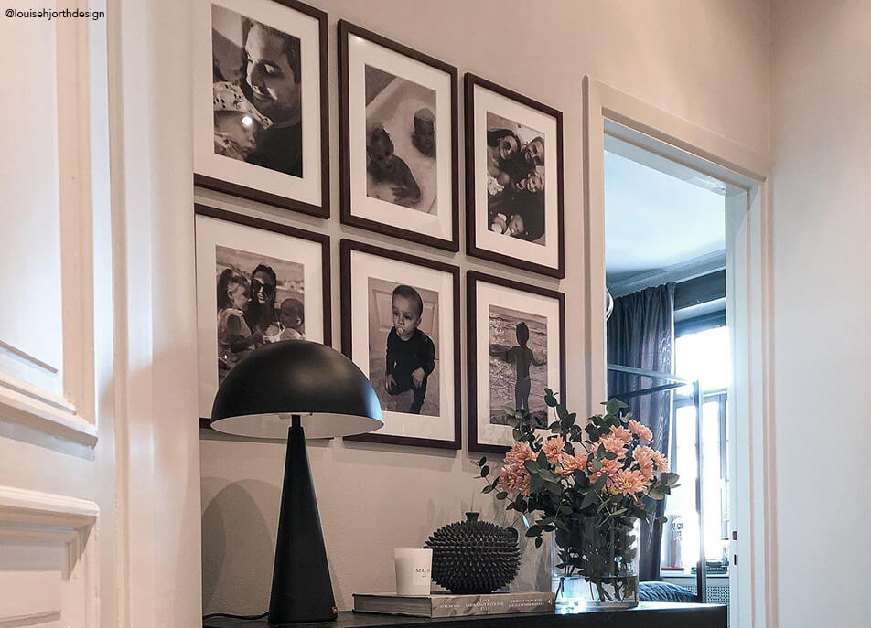 Fotografijų siena prie prieškambario, juodai baltos nuotraukos tamsiuose mediniuose rėmuose