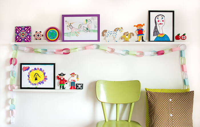 Stilingi balti paveikslų lentynėlės, dekoruoti spalvotais vaikų piešiniais ir kitais gražiais daiktais Balta paveikslinė lentelė vaikų kambaryje
